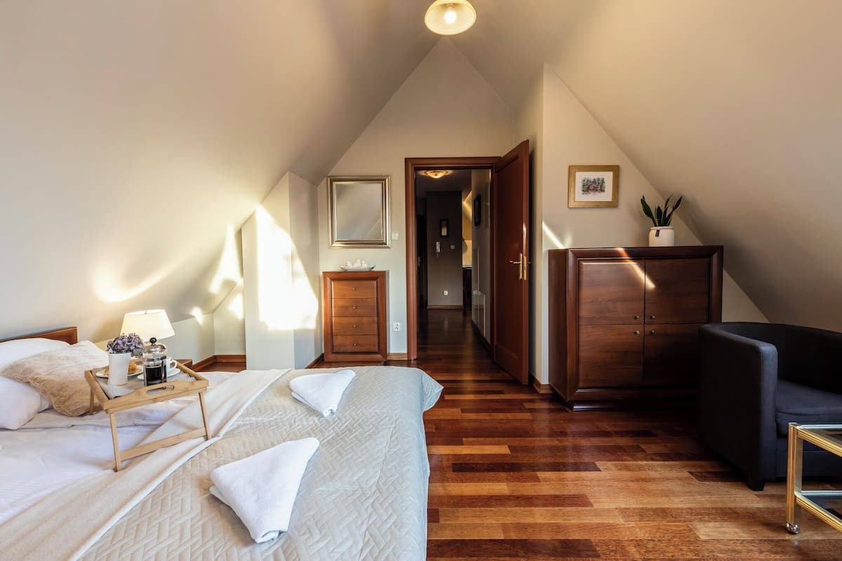 Un dormitorio con piso de madera, una cama con ropa de cama blanca, una mesita de noche con lámpara, un aparador y un sillón de color oscuro.