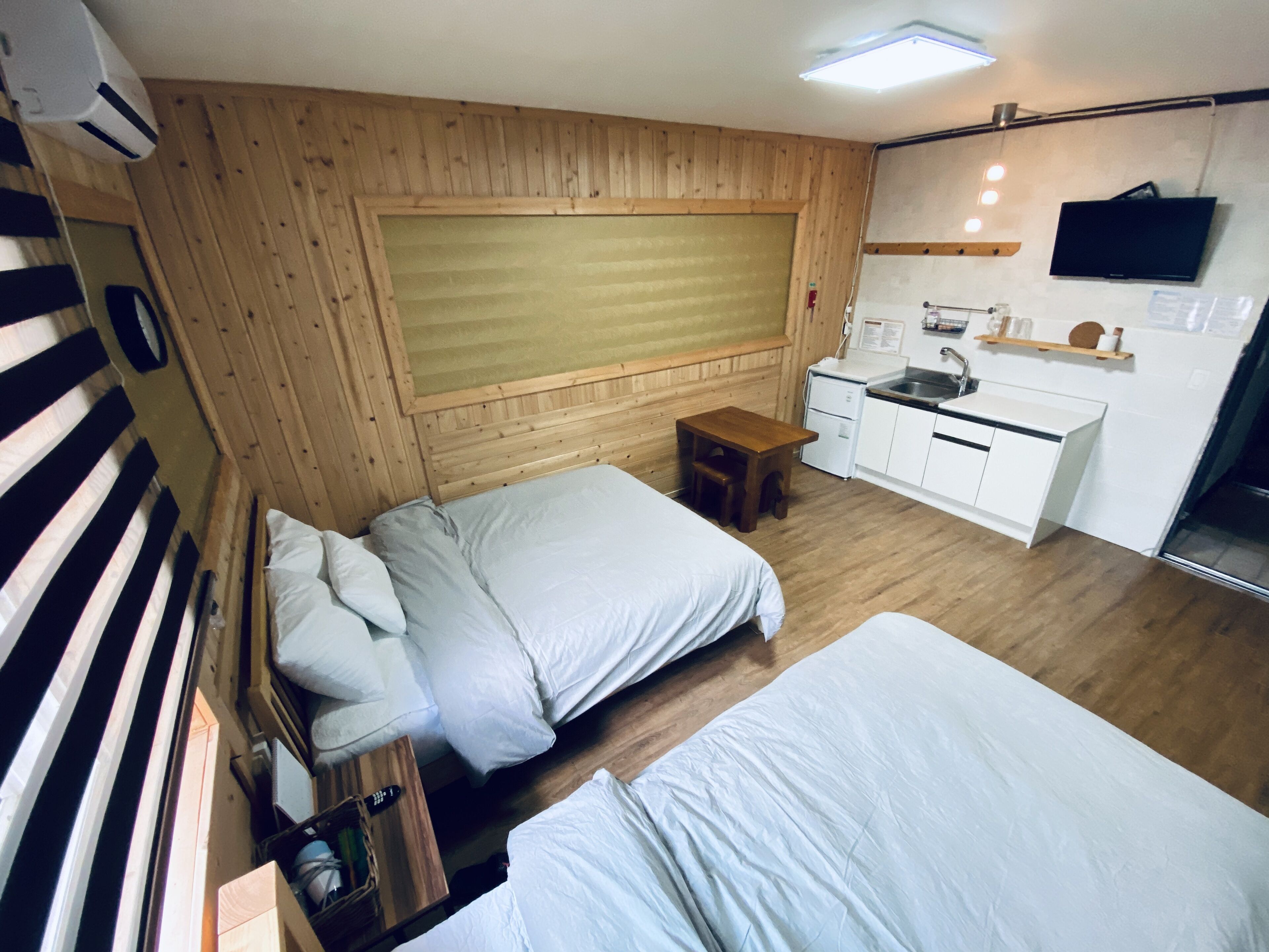 A compact room with two beds, a small wooden table, a kitchenette, and a mounted TV.