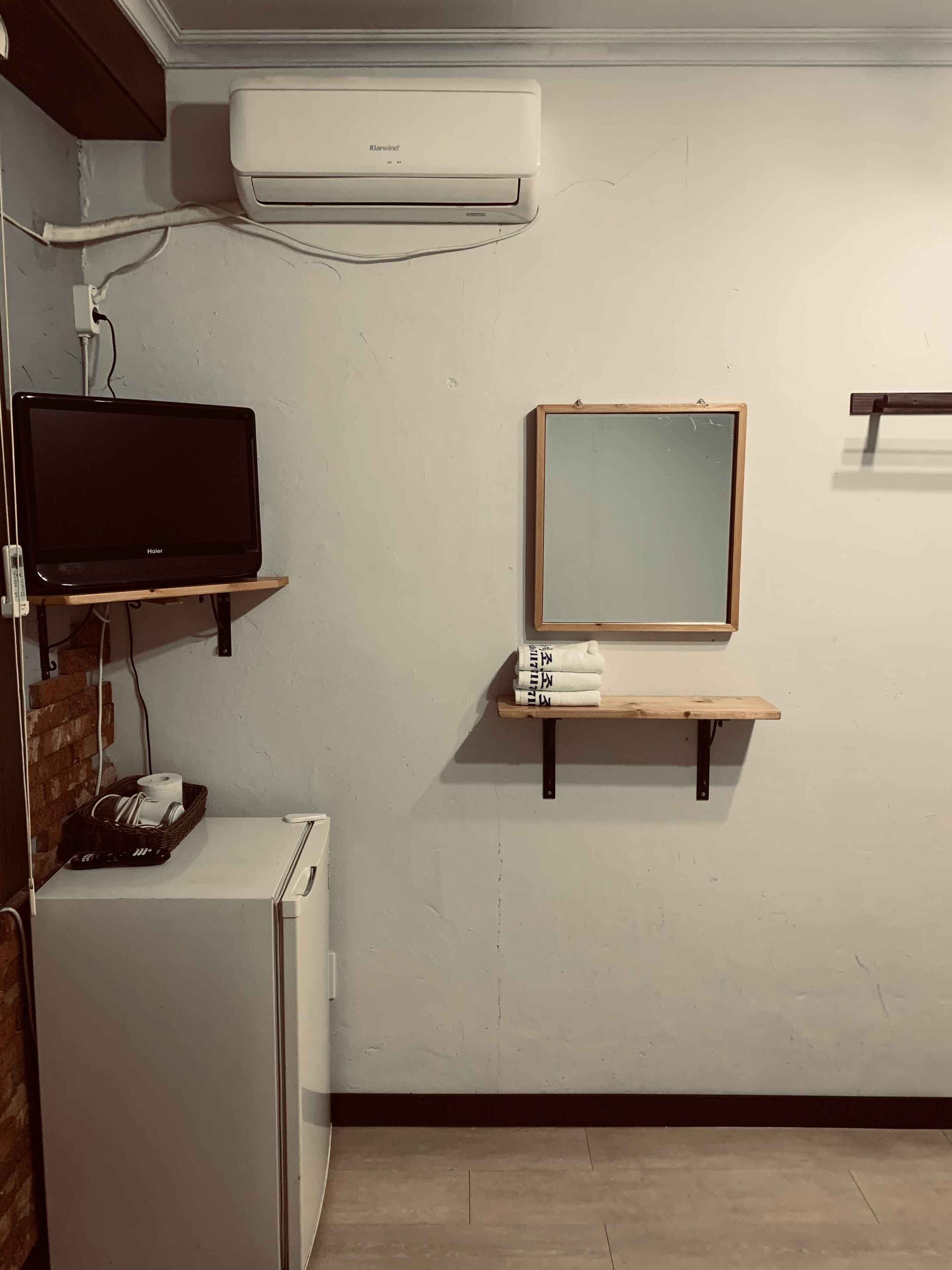 A compact room with a white air conditioner, a black television on a shelf, a small refrigerator, a wooden shelf with towels, and a mirror on the wall.