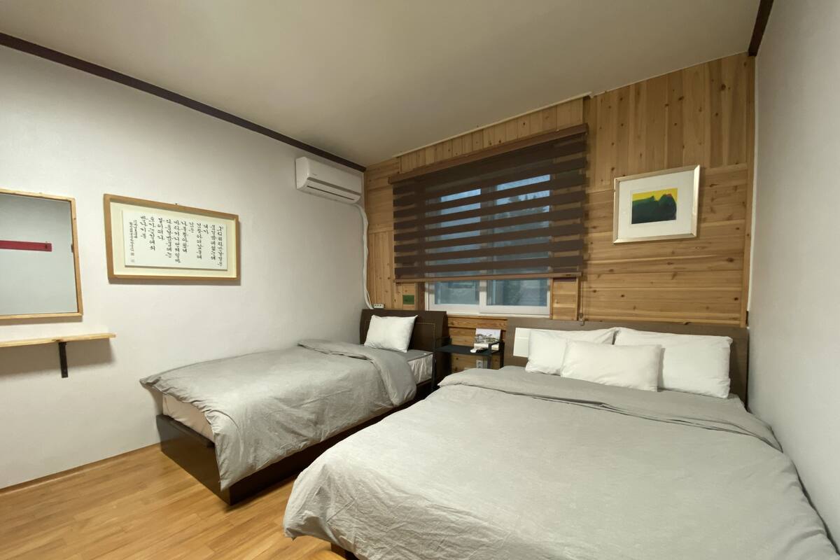 A bedroom with two beds, wooden paneling, a window with blinds, and a wall-mounted air conditioner.
