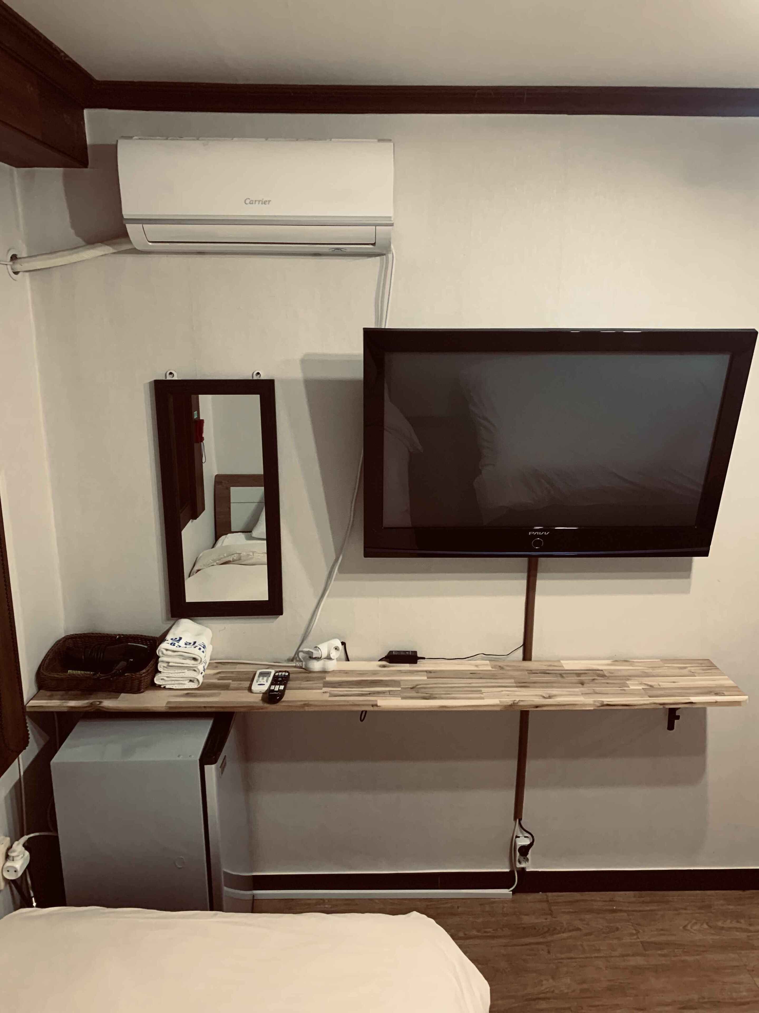 A hotel room with a bed, a TV mounted on a wooden shelf, a mirror, and an air conditioning unit.
