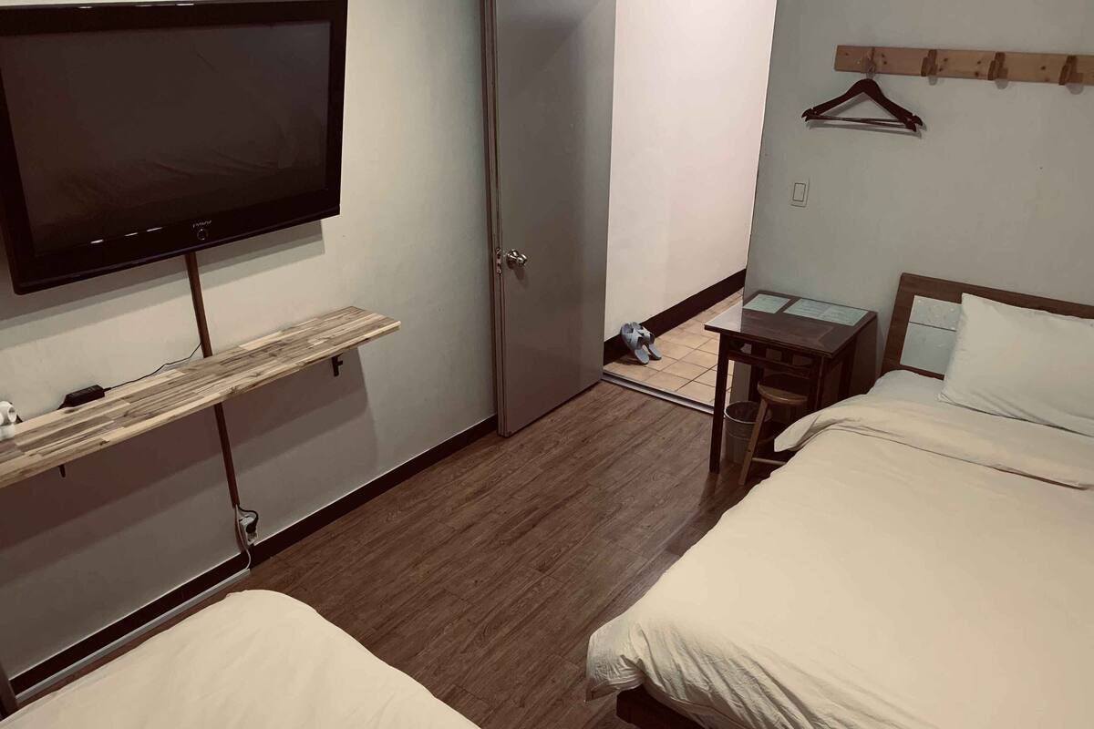 A hotel room with two beds, a TV mounted on the wall, a wooden shelf, a small table with a chair, and a coat rack.