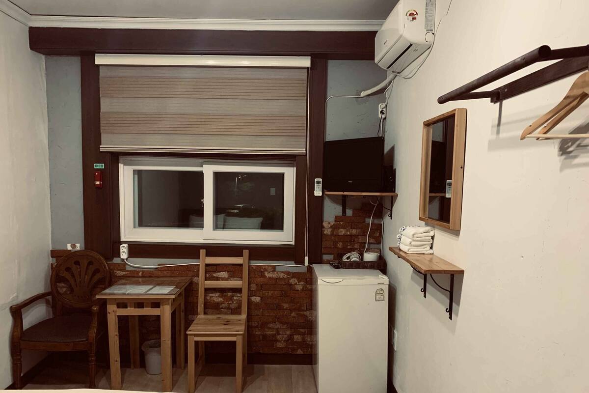 A compact room with a small table, two chairs, a refrigerator, a window with blinds, a wall-mounted TV, and a wooden shelf with towels.