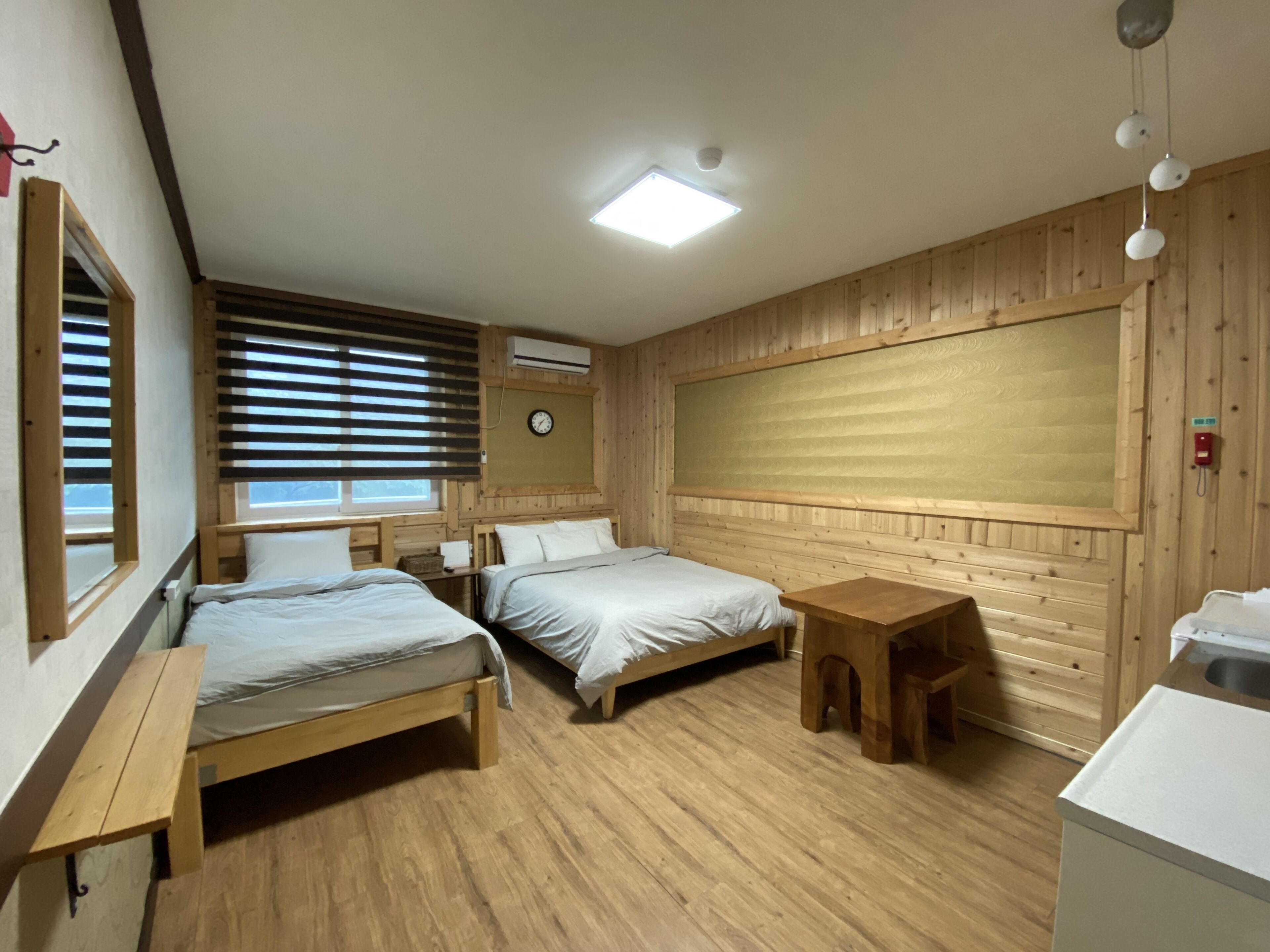 A room with two wooden beds, a small wooden table, a white sink, and a window with blinds.