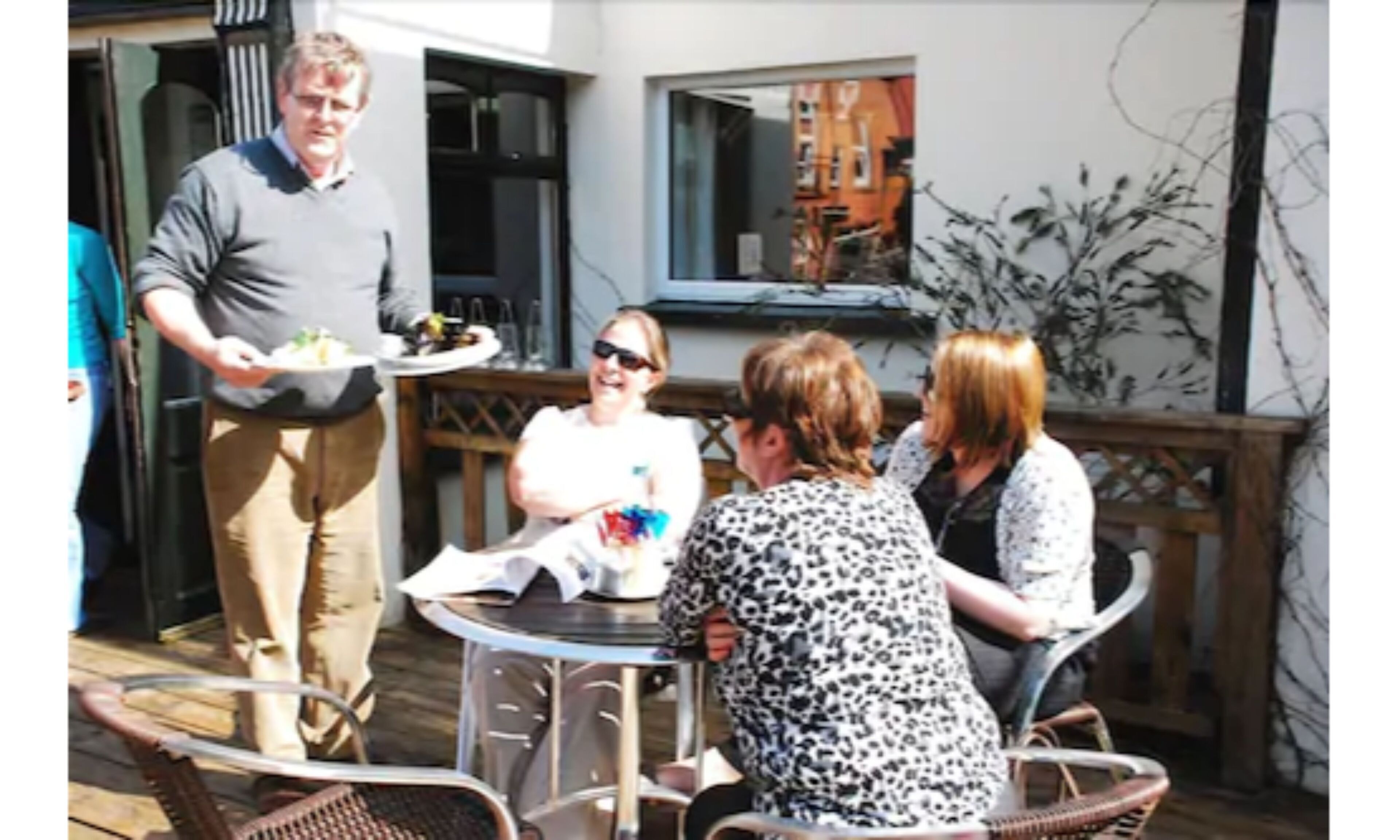 Un grupo de personas sentadas en una mesa al aire libre sobre una terraza de madera, con un edificio de fondo.