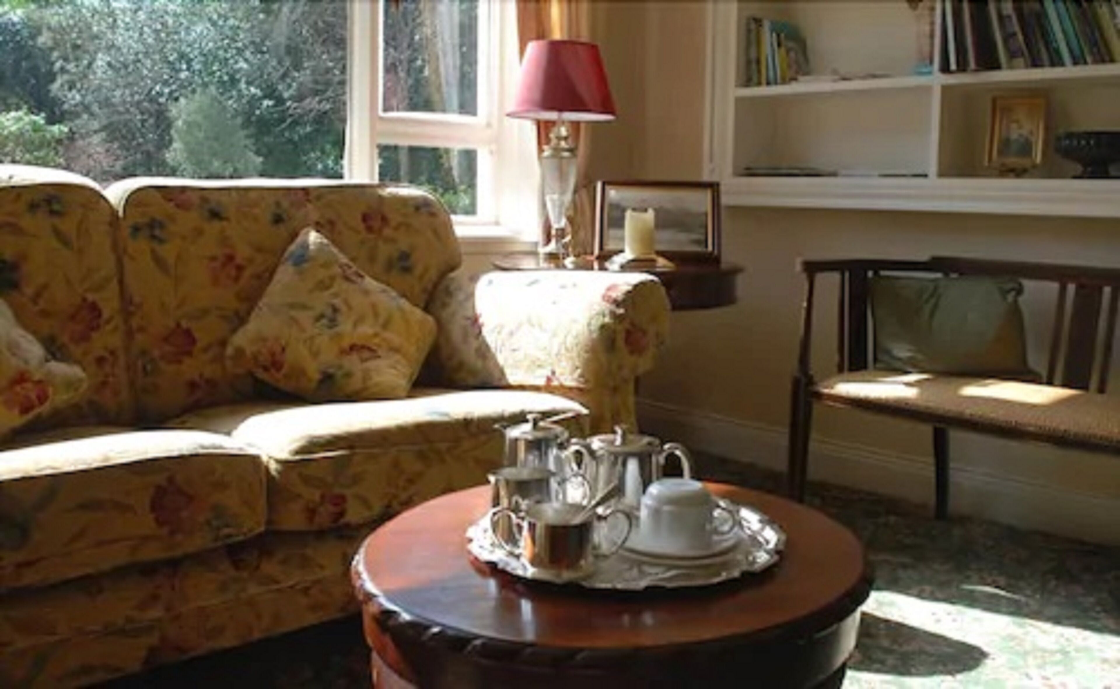 A cozy living room with a floral sofa, a wooden coffee table with tea set, a red lamp, and a bookshelf.