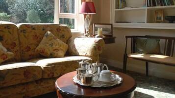 A cozy living room with a floral sofa, a wooden coffee table with tea set, a red lamp, and a bookshelf.