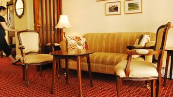 Habitación de hotel con alfombra roja estampada, un sofá, dos sillones, una mesita de noche con una lámpara y un espejo en la pared.