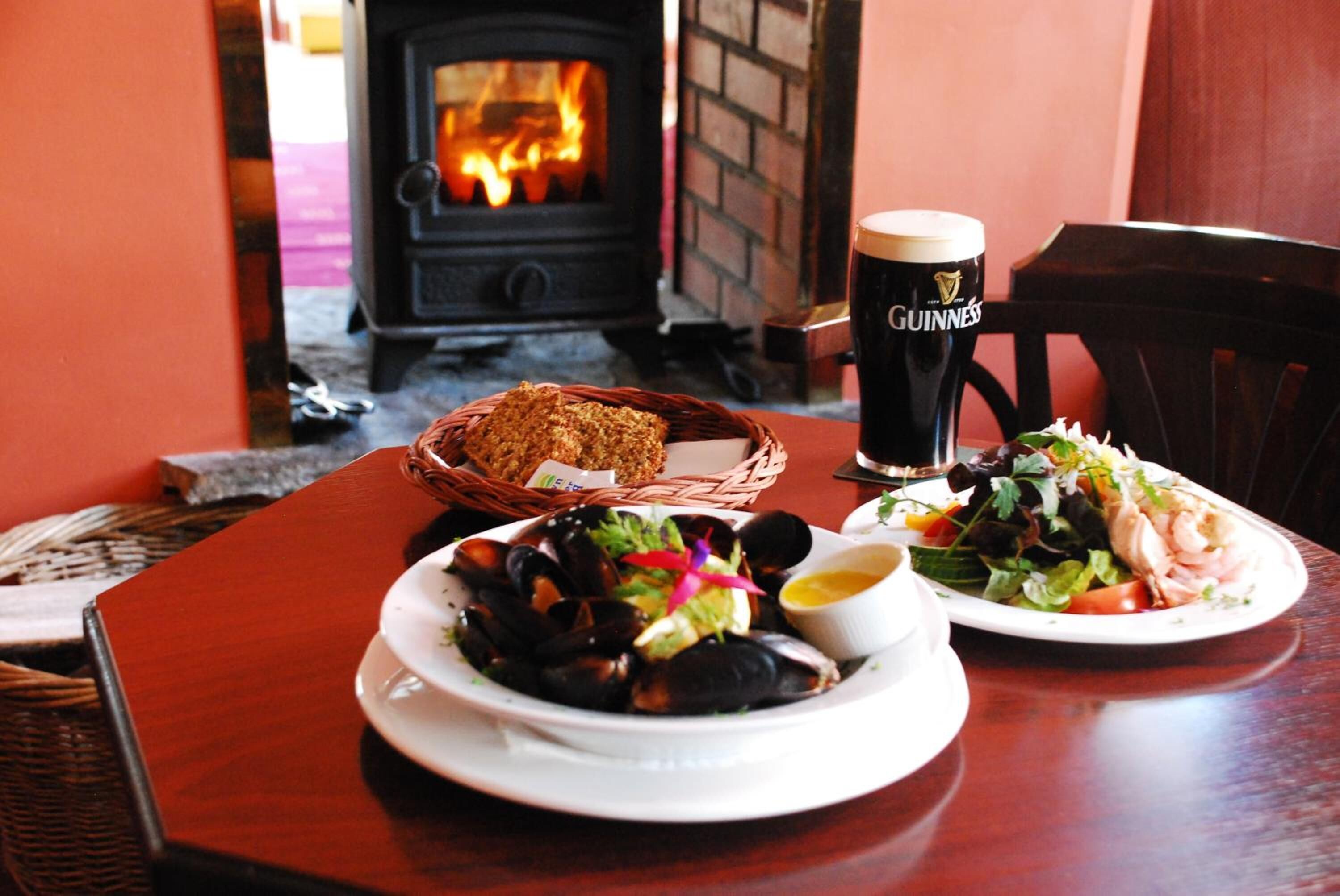 A table with a plate of mussels, a salad, and a loaf of bread, a glass of Guinness beer, and a fireplace in the background.