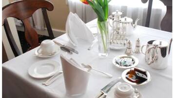 Mesa dispuesta para comer, con mantel blanco, cubiertos, platos, una jarra, un florero con flores y una tetera.
