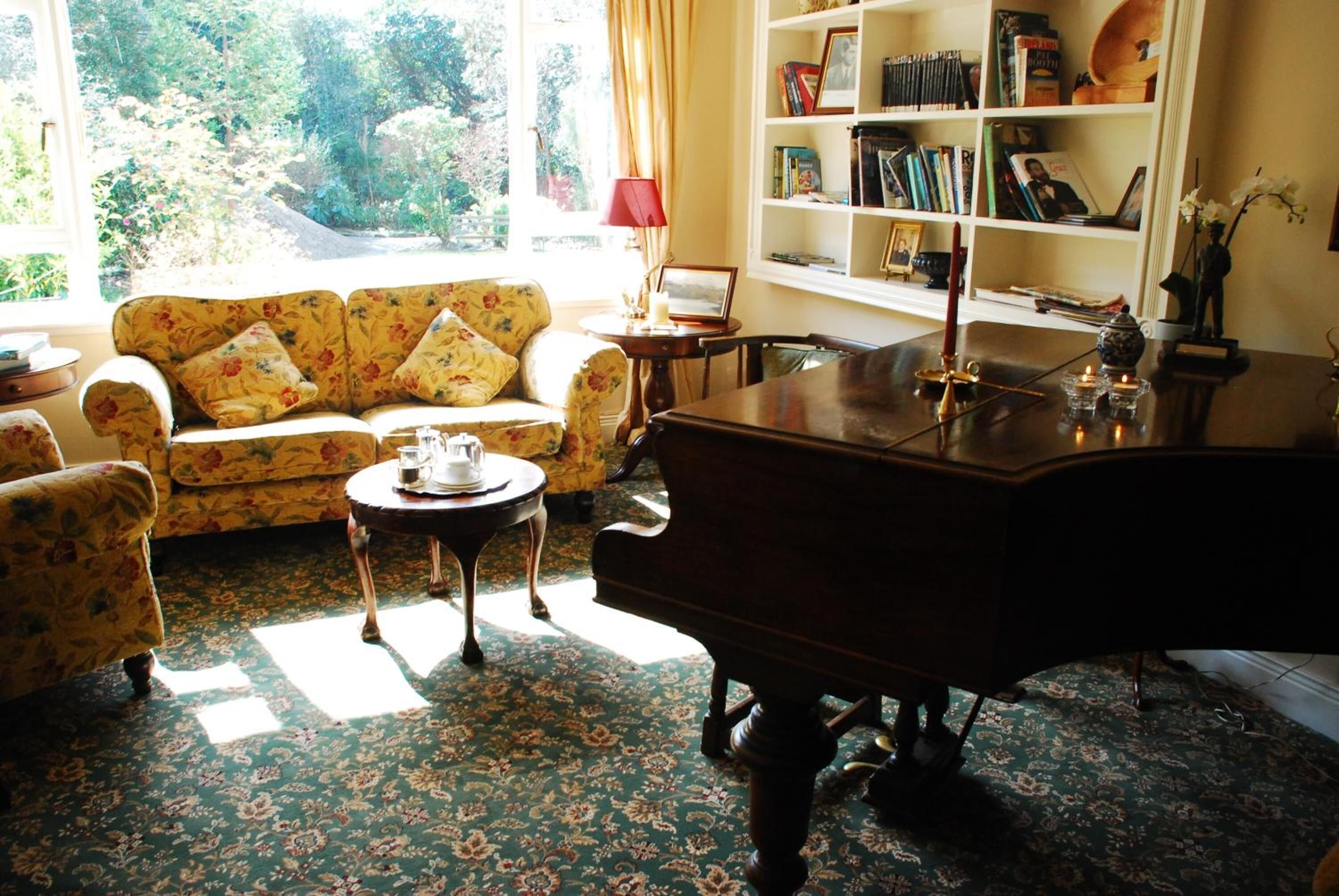 Una sala de estar con un sofá floral, una mesa de centro de madera, un piano de cola y una estantería llena de libros y objetos decorativos.