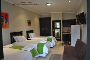 Habitación de hotel con tres camas, kitchenette, televisor y obras de arte en las paredes.