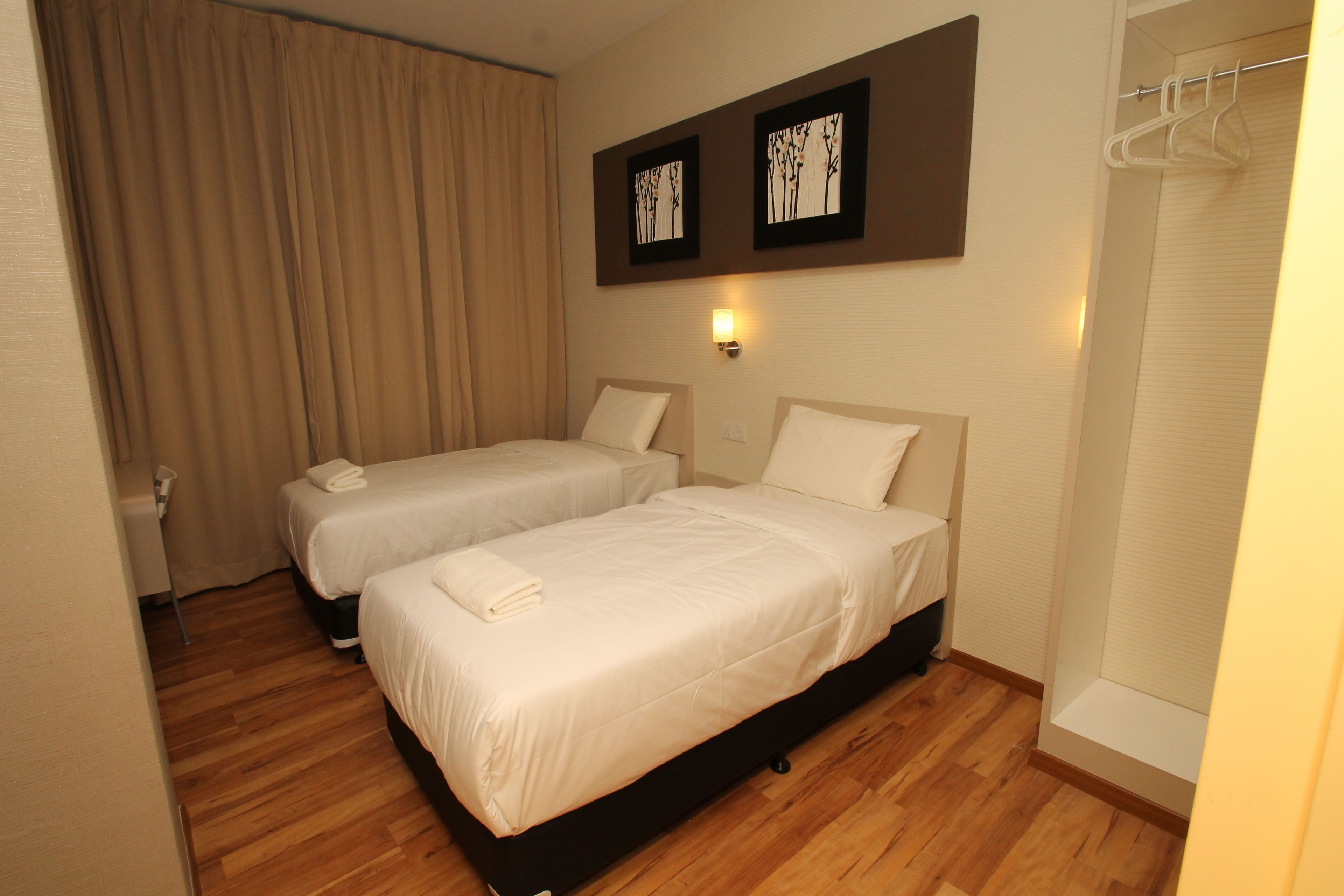 Habitación de hotel con dos camas individuales, suelo de madera, un ventanal grande con cortinas y dos marcos con cuadros en la pared.