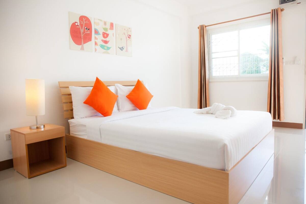 A neatly made bed with white and orange pillows, a bedside lamp, and a window with curtains.