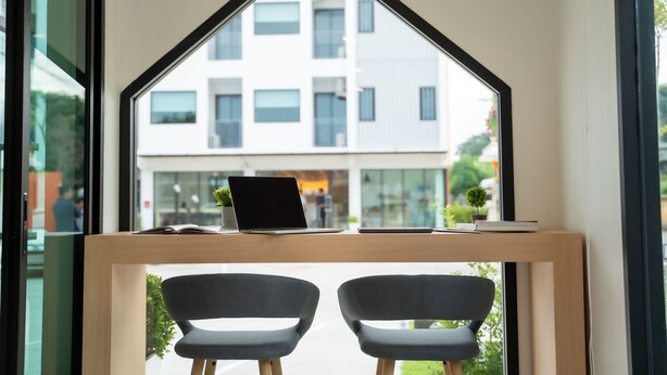 A modern workspace with a laptop, a book, and a small plant on a wooden desk. Two chairs are positioned in front of a window with a view of a building. - Meesuk ChiangRai Hotel, SHA Certified (Chiang Rai)