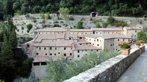 Un bâtiment historique en pierre, doté de toits couverts de tuiles en terre cuite, entouré d’une végétation luxuriante et d’un mur de pierre.