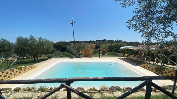 Une piscine rectangulaire, avec une clôture en bois au premier plan, entourée de verdure et un petit bâtiment en arrière-plan.