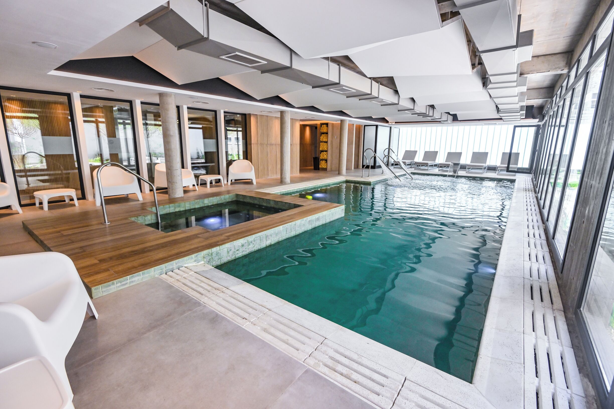 Ein Innenpool mit Holzterrasse, Liegestühlen und einer Glaswand mit Blick ins Freie.