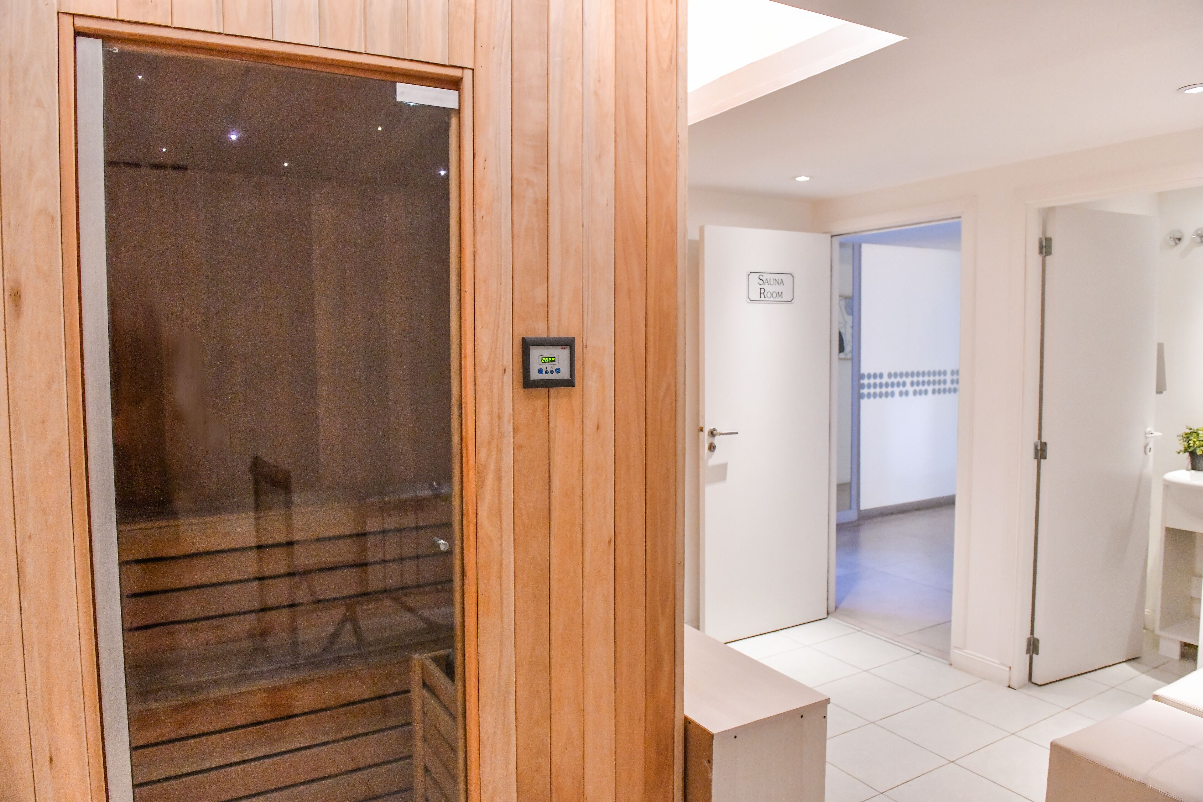Un sauna en bois avec une porte en verre, un mur blanc portant l’inscription « Sauna », et un sol carrelé menant à une salle de bain équipée d’un lavabo.