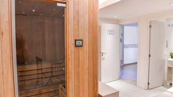 Un sauna en bois avec une porte en verre, un mur blanc portant l’inscription « Sauna », et un sol carrelé menant à une salle de bain équipée d’un lavabo.