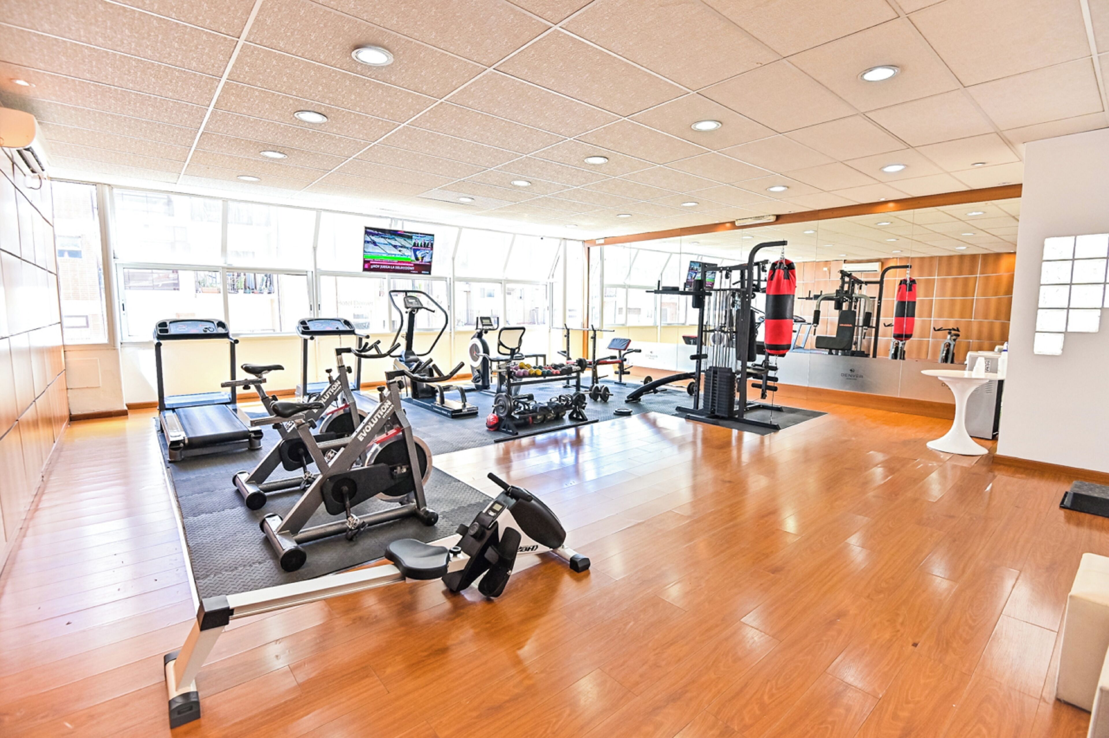 A spacious gym with various exercise equipment, large windows, and a wooden floor.