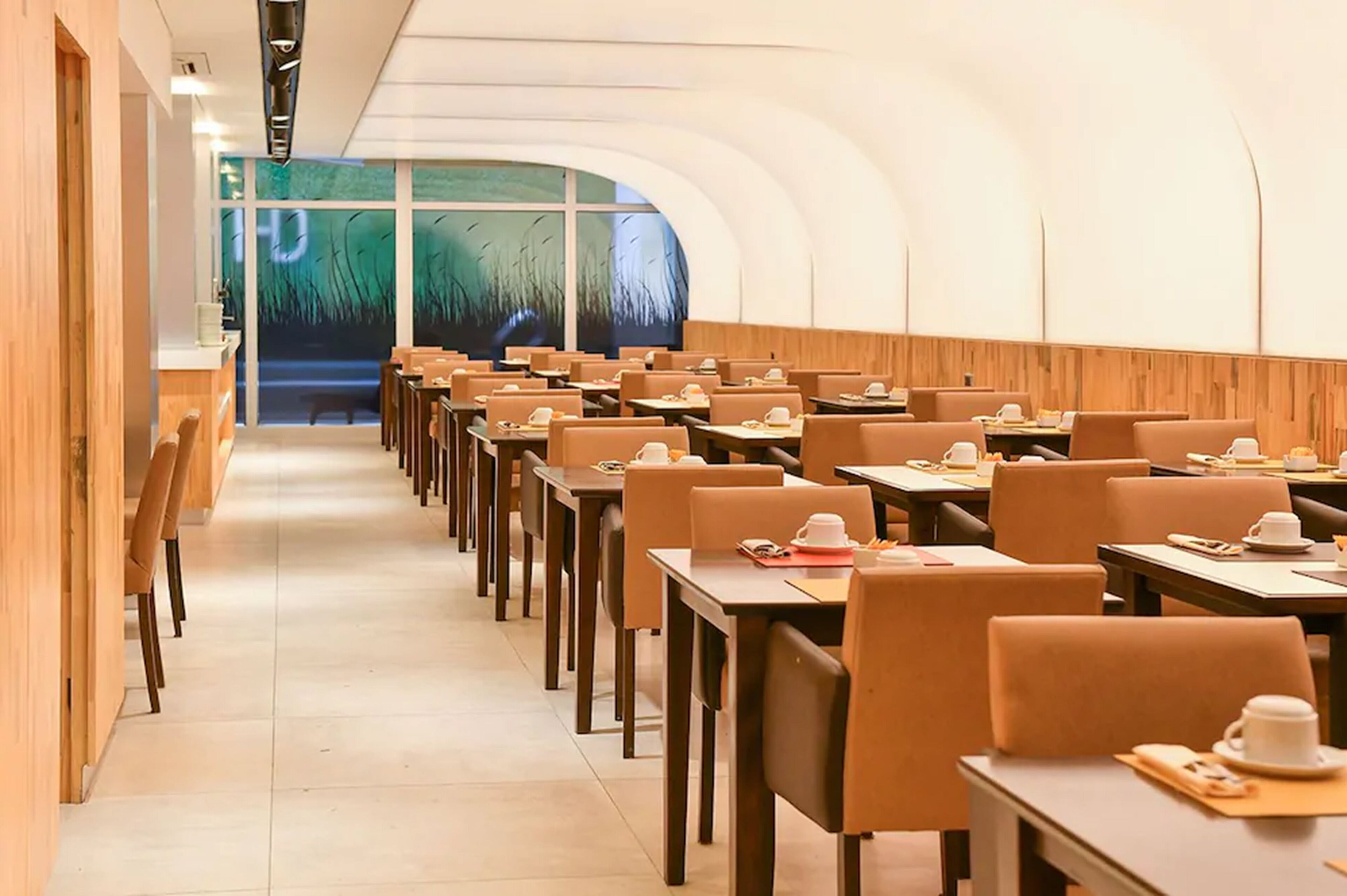 Ein modernes Restaurant mit langen Tischen und Stühlen, einem hölzernen Empfangstresen und einem großen Fenster mit Blick auf hohes Gras.
