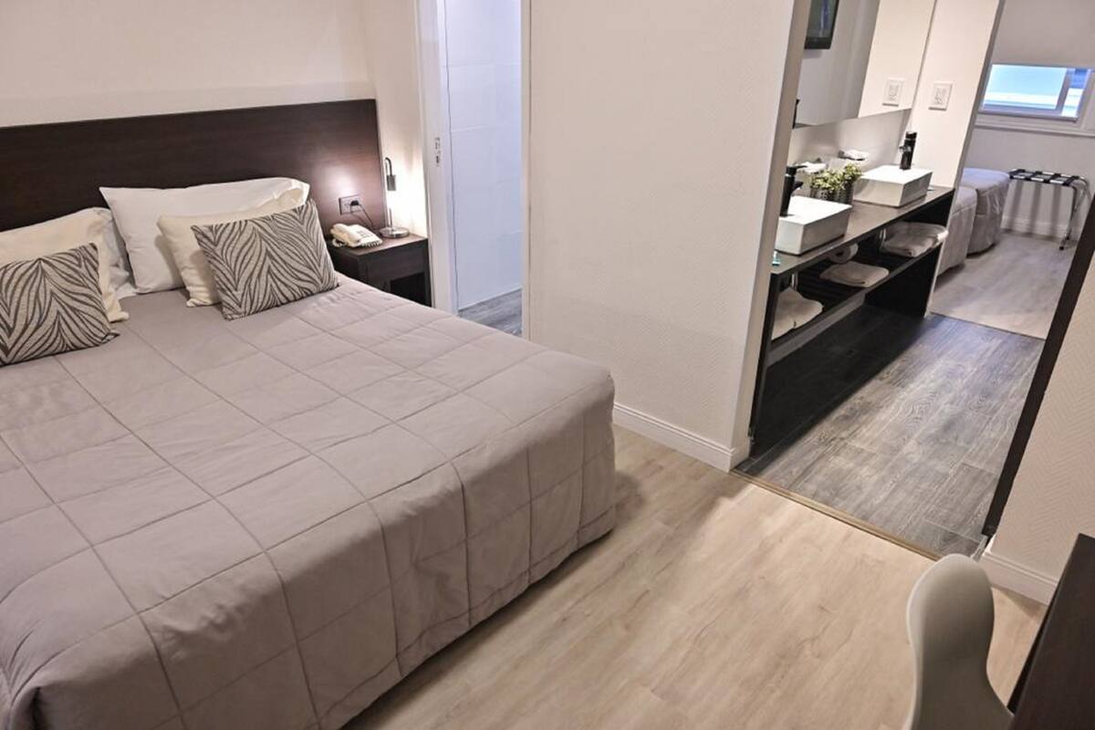 A modern hotel room with a large bed, a bedside table with a lamp, a bathroom with a sink and mirror, and a view of the hallway.