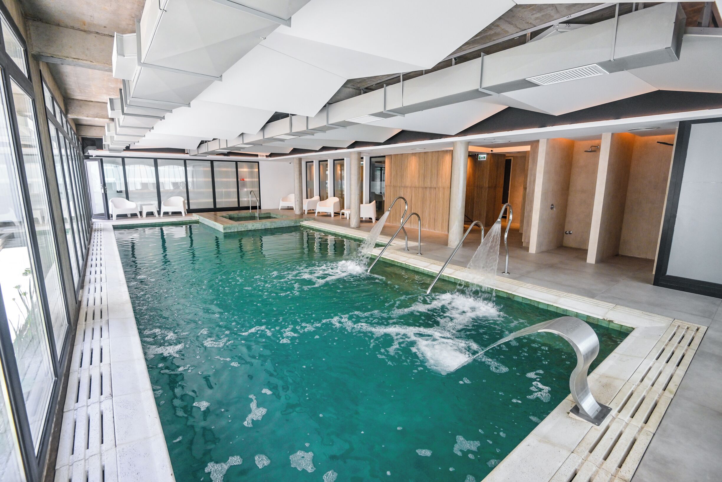 A modern indoor swimming pool with clear water, surrounded by white chairs and a wooden wall.