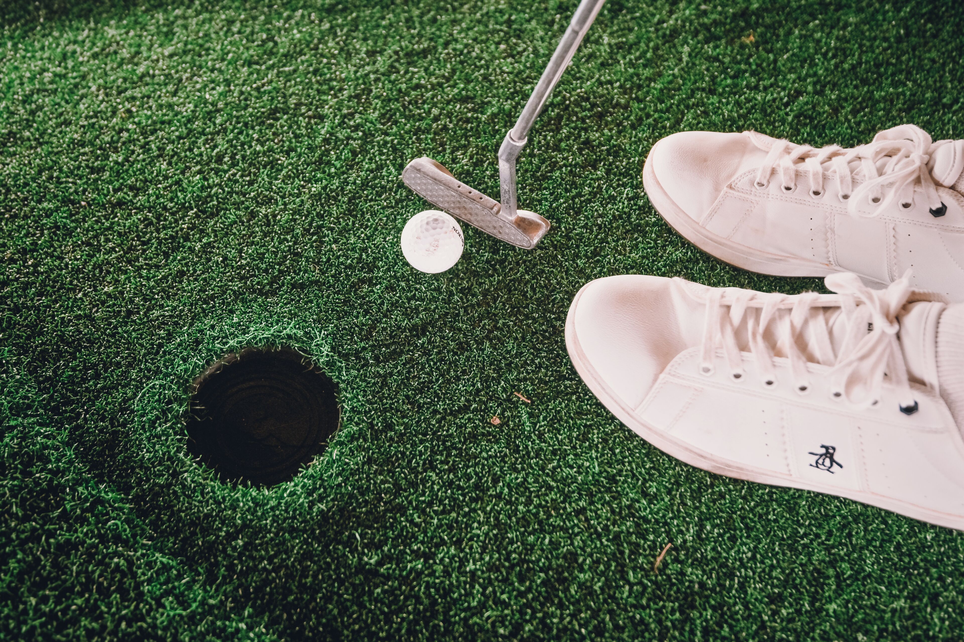 A pair of white sneakers and a golf club on a green surface with a hole.