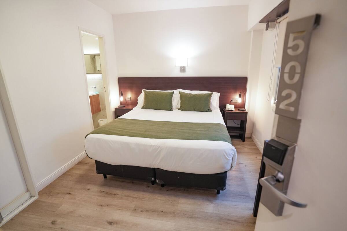 A hotel room with a large bed, a nightstand with a lamp, a phone, and a wall-mounted number sign.