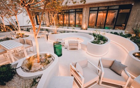 An outdoor seating area with modern white furniture, surrounded by planters and trees, in front of a building with large windows.