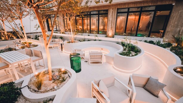 An outdoor seating area with modern white furniture, surrounded by planters and trees, in front of a building with large windows. - Hotel Denver (Mar del Plata)
