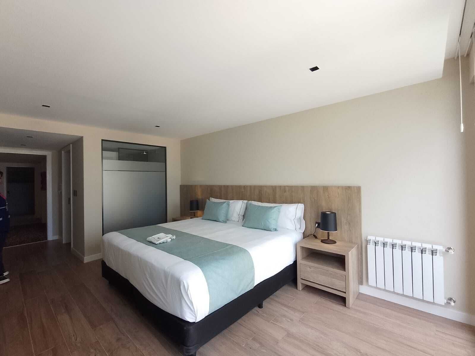 Una habitación de hotel moderna con una cama grande, cabecera de madera, mesita de noche con lámpara y radiador.