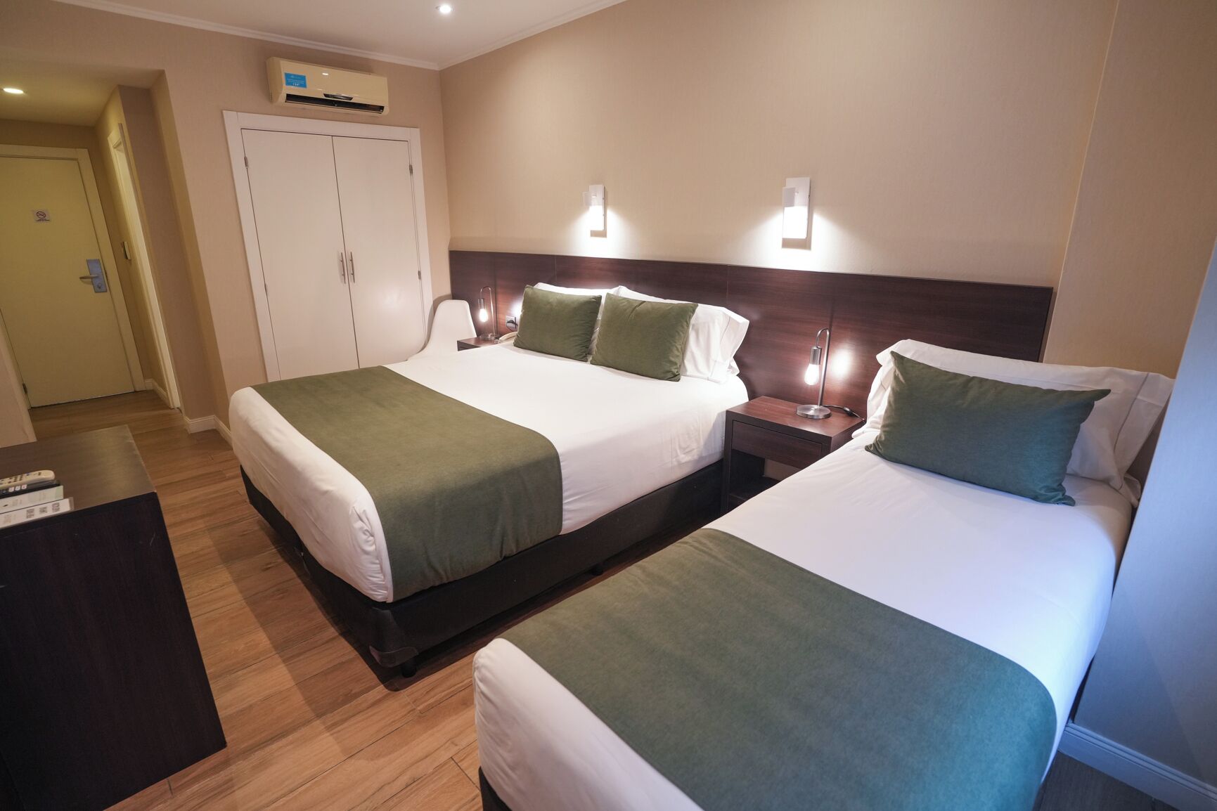 Habitación de hotel con dos camas, un armario, un escritorio y una mesita de noche con lámpara.