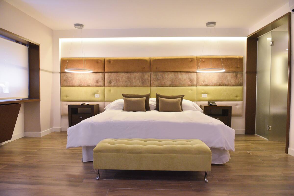 A modern hotel room with a large bed, a bedside table, a bench, and a headboard with built-in lighting.
