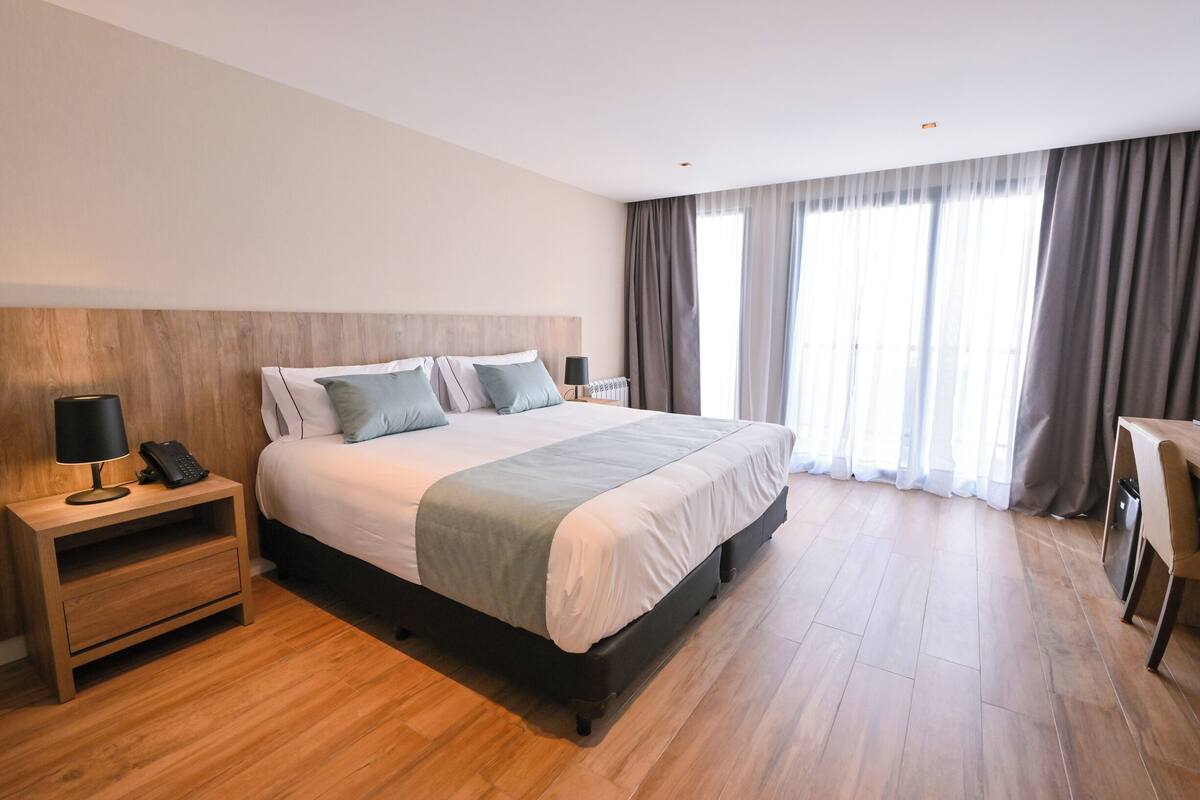 A modern hotel room with a wooden headboard, a large bed with white and grey bedding, a bedside table with a lamp and phone, and a view of the city through large windows.