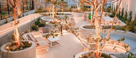 An outdoor seating area with white chairs and tables, surrounded by planters and trees, with a building in the background.