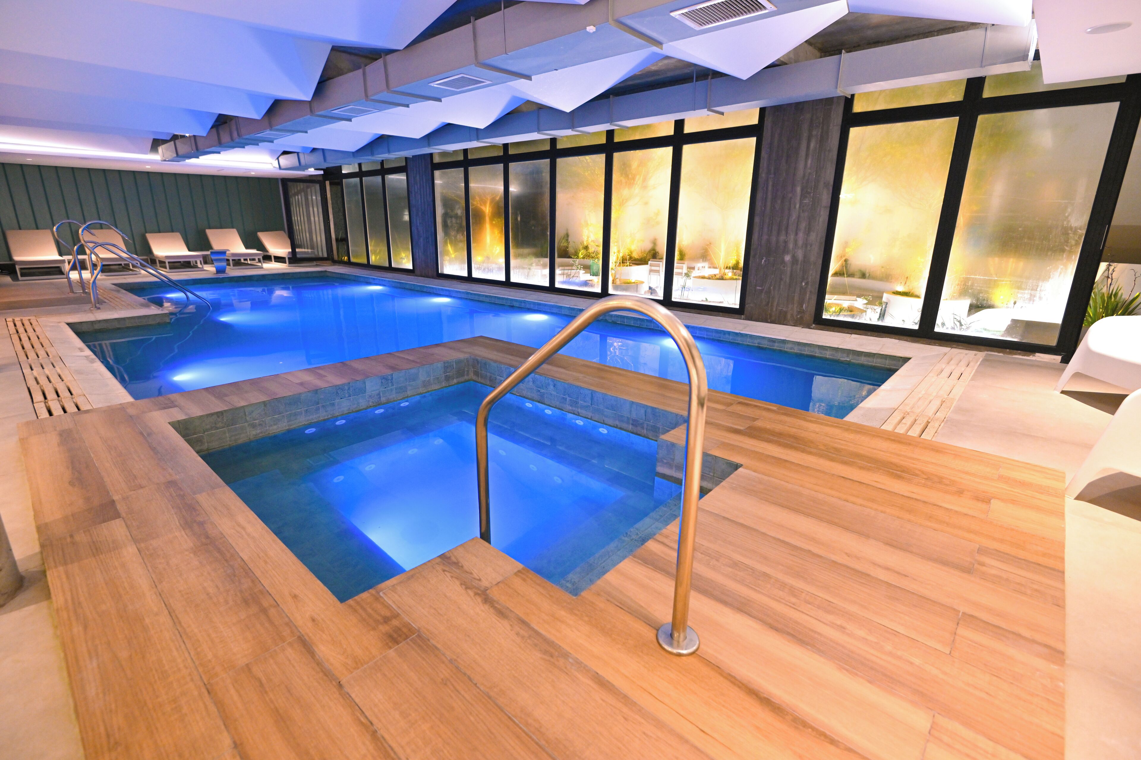 Une piscine intérieure dotée d’une terrasse en bois, de chaises longues et de grandes fenêtres offrant une vue sur l’extérieur.