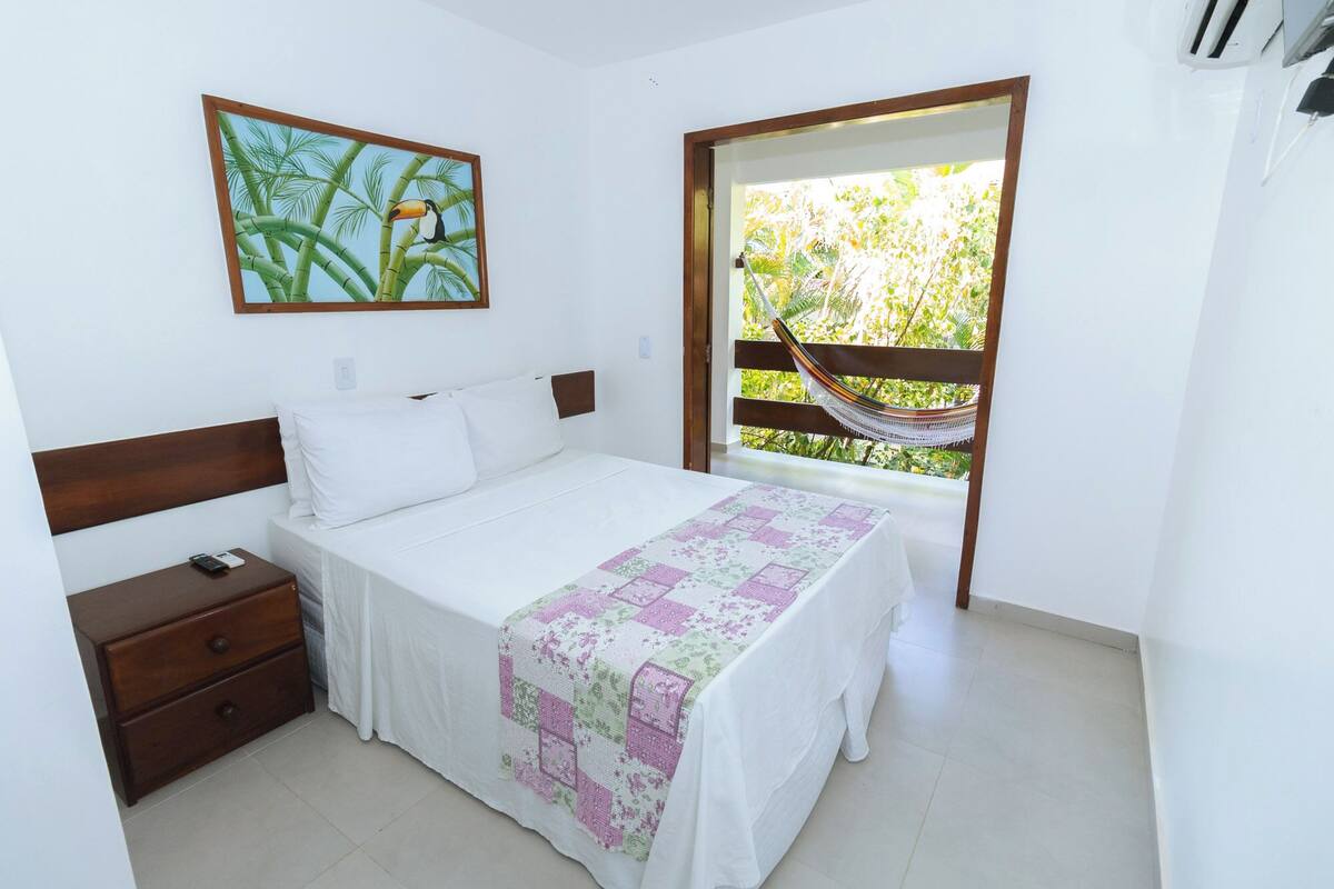 A bedroom with a bed, a nightstand, a wooden headboard, a hammock, and a window with a view of greenery.