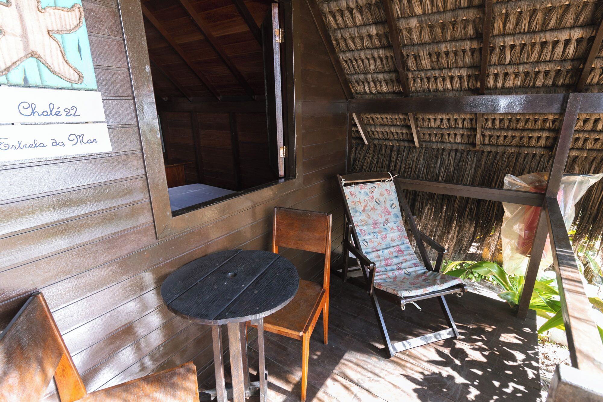 A wooden room with a thatched roof, a small table, and two chairs. A sign on the wall reads "Chalet 22 Estrela da Mar".