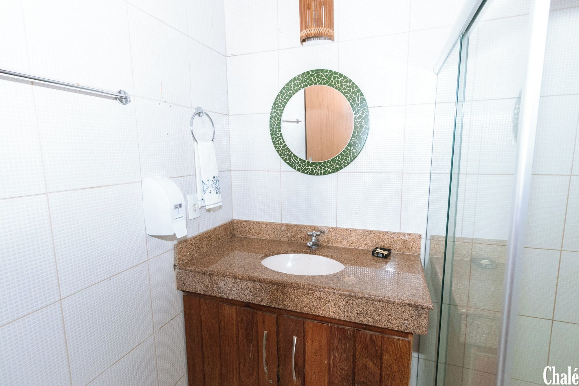 A bathroom with a granite countertop sink, wooden cabinets, a towel dispenser, and a glass shower enclosure.