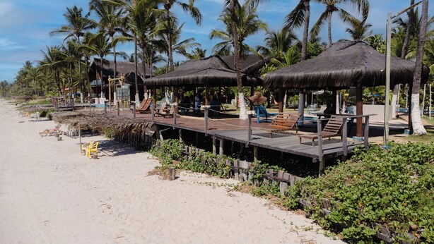 Een resort aan het strand met rieten hutten, houten terraszitplaatsen en palmbomen.