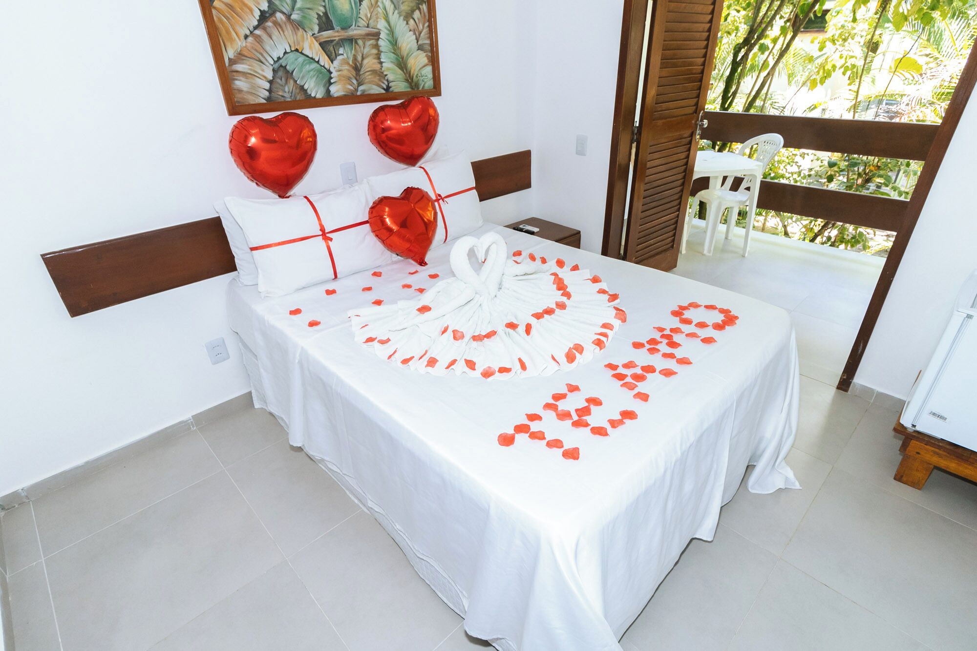 A neatly arranged bed with a white bedspread, red heart-shaped balloons, and a heart-shaped arrangement of rose petals.