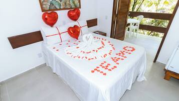 A neatly arranged bed with a white bedspread, red heart-shaped balloons, and a heart-shaped arrangement of rose petals.