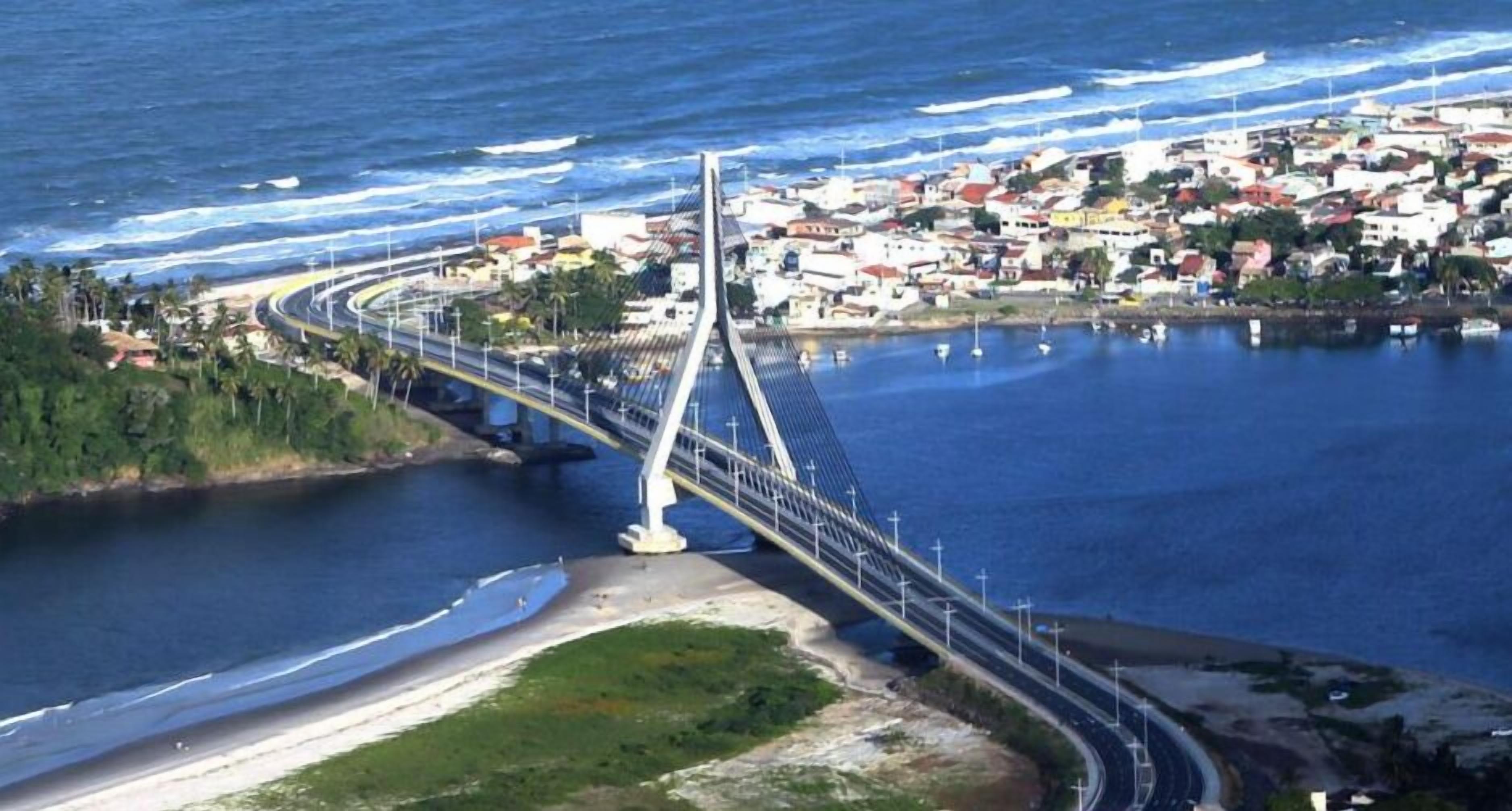 Un puente costero con una pasarela peatonal, que une una zona muy poblada con una playa.