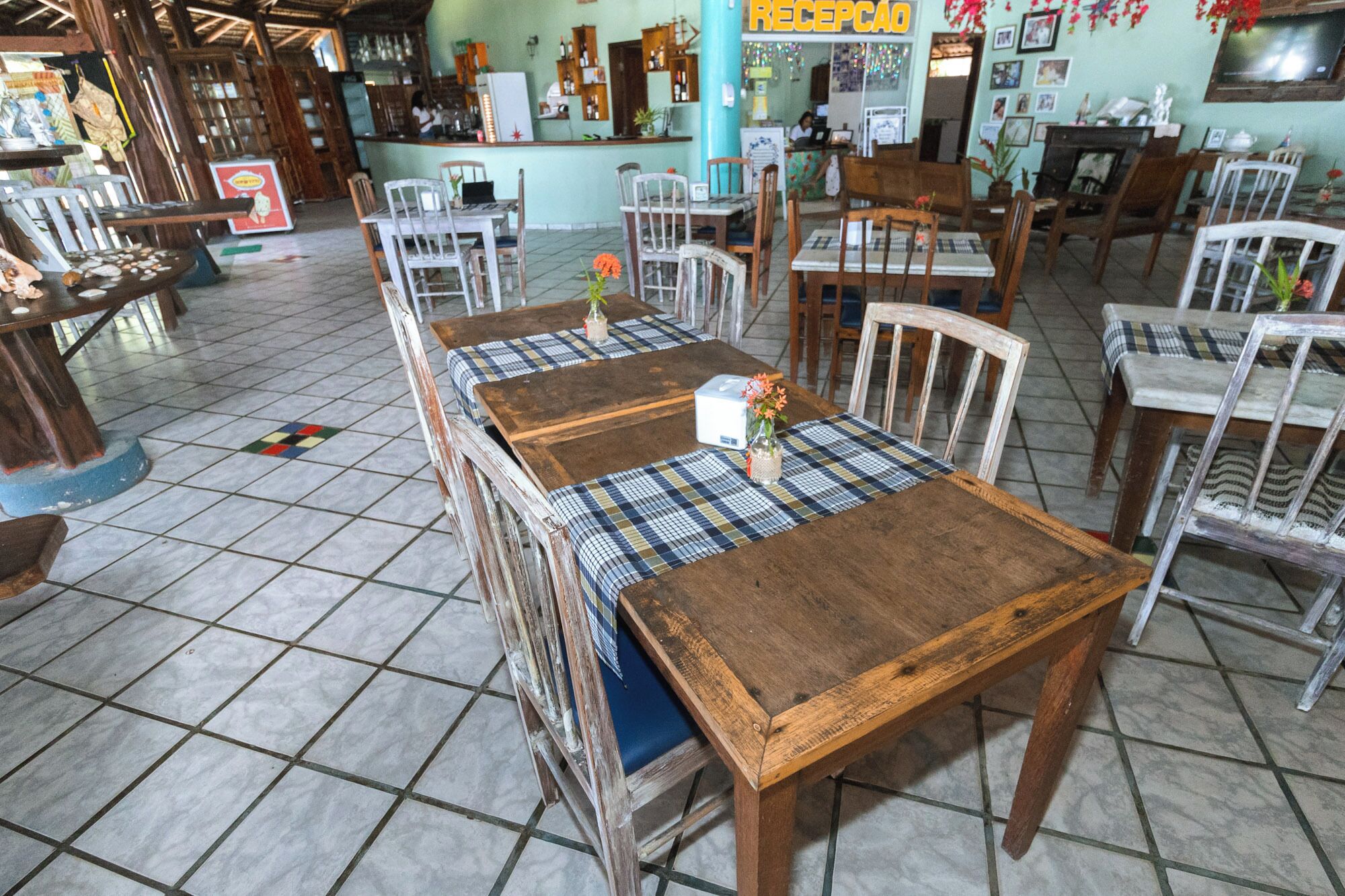A restaurant interior with wooden tables and chairs, a tiled floor, and a reception area in the background.