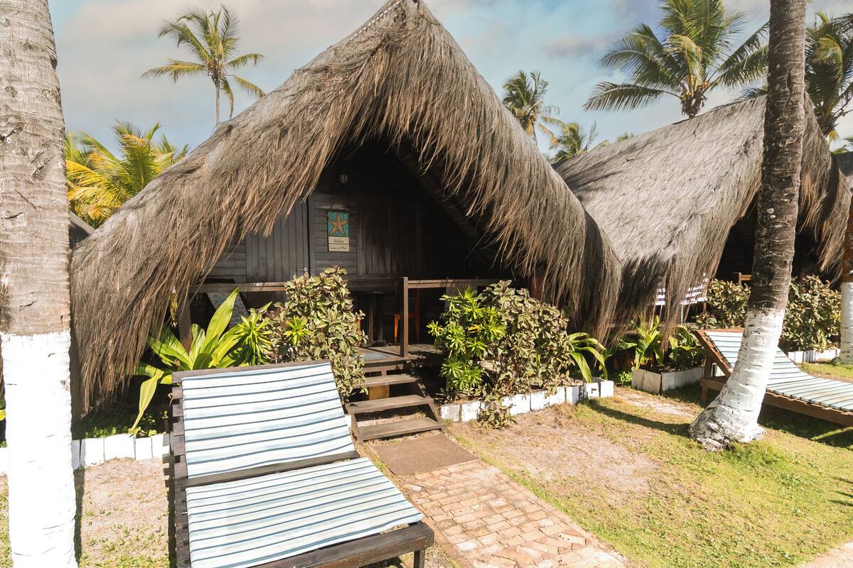 A thatched-roof structure with a wooden door, surrounded by palm trees and a paved walkway leading to striped lounge chairs.
