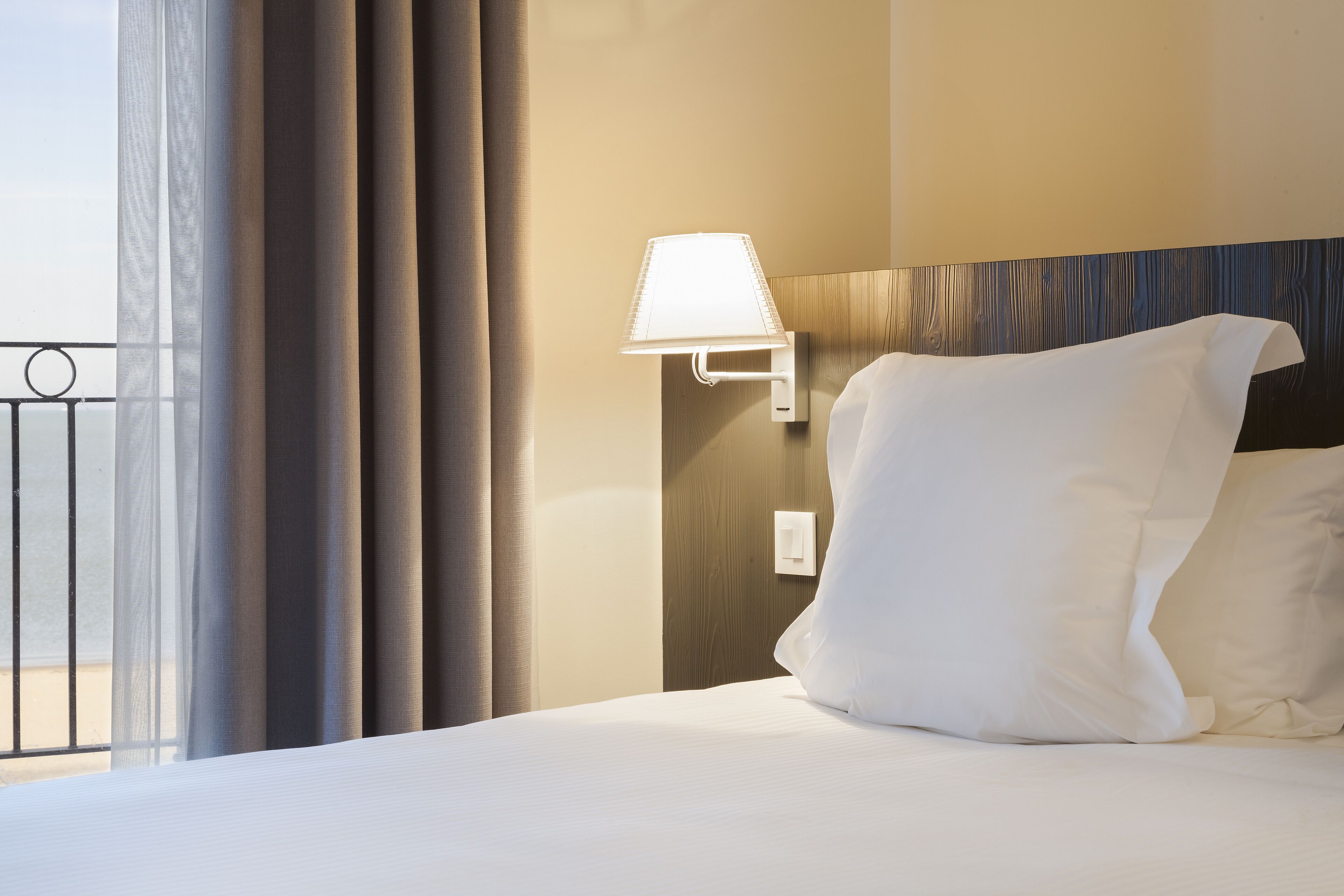 Une chambre d’hôtel avec un lit, une lampe fixée au mur et une vue sur l’extérieur par une fenêtre munie de rideaux.