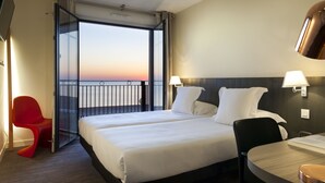 A hotel room with two beds, a red chair, a table, and a view of the sunset.