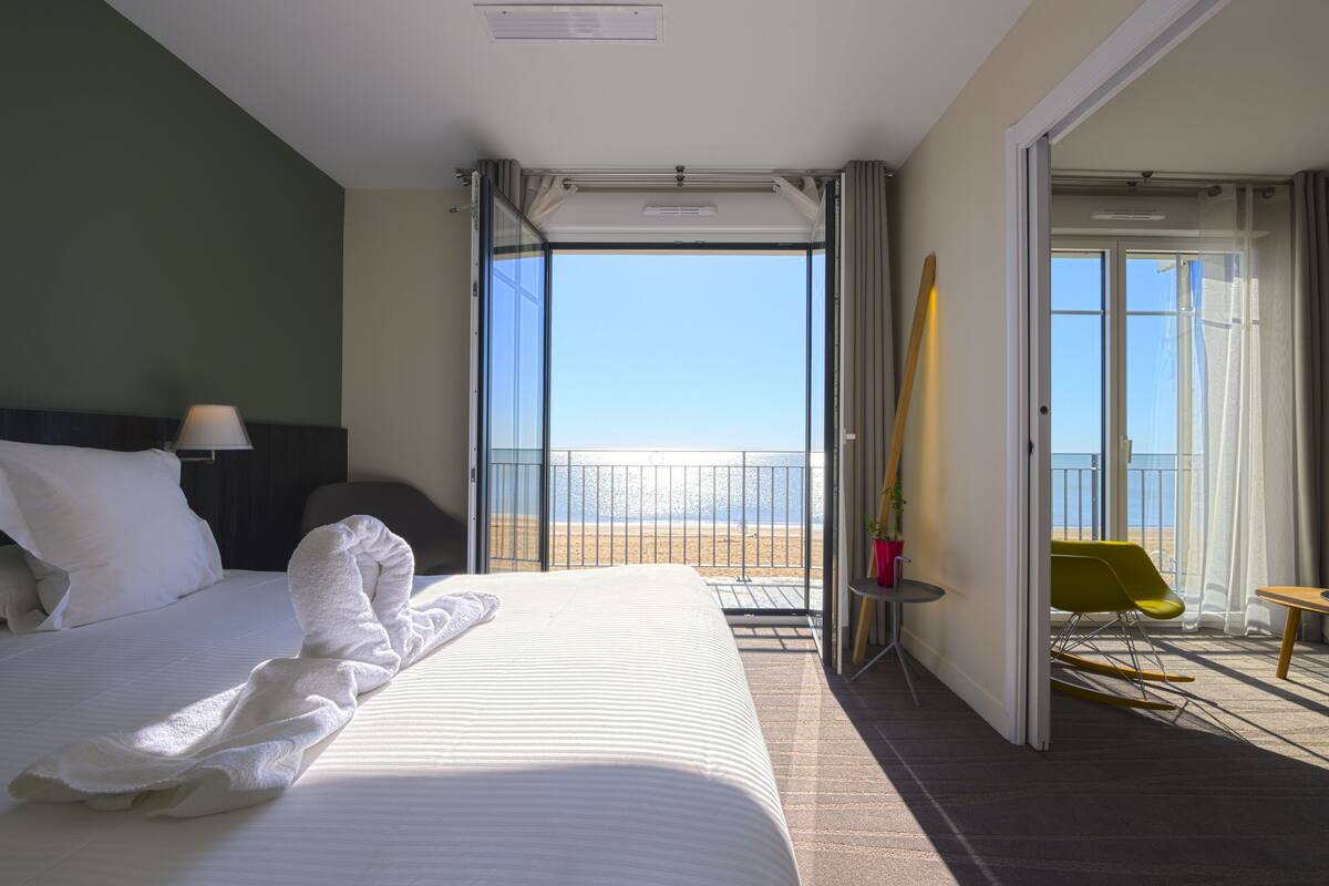 Una habitación de hotel con cama, balcón y vista a la playa, además de una silla.